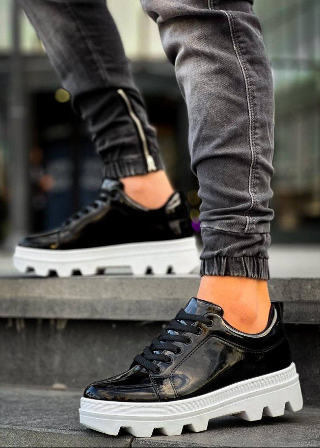 Black glossy sneakers with white soles worn by a person on a concrete surface.